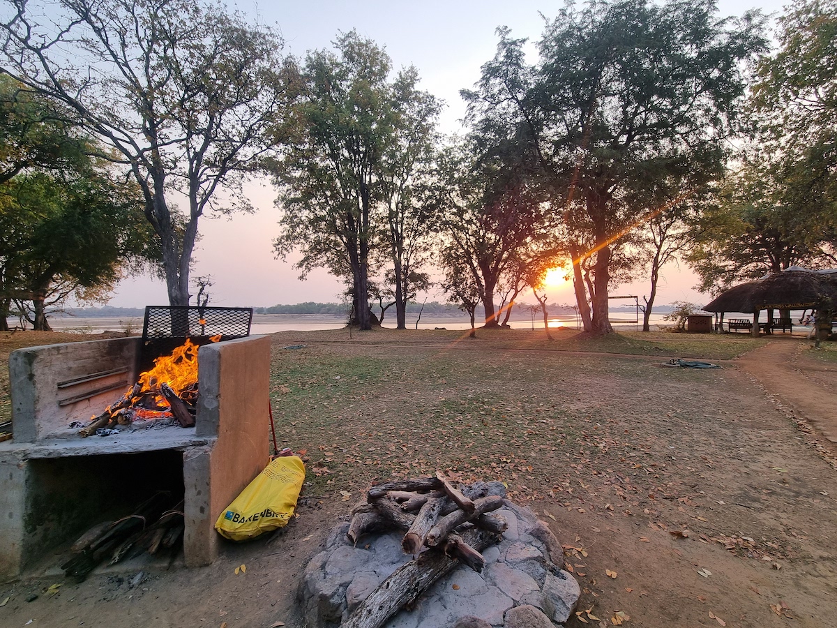 A braai fire at sunset beside a river and thatched lapa