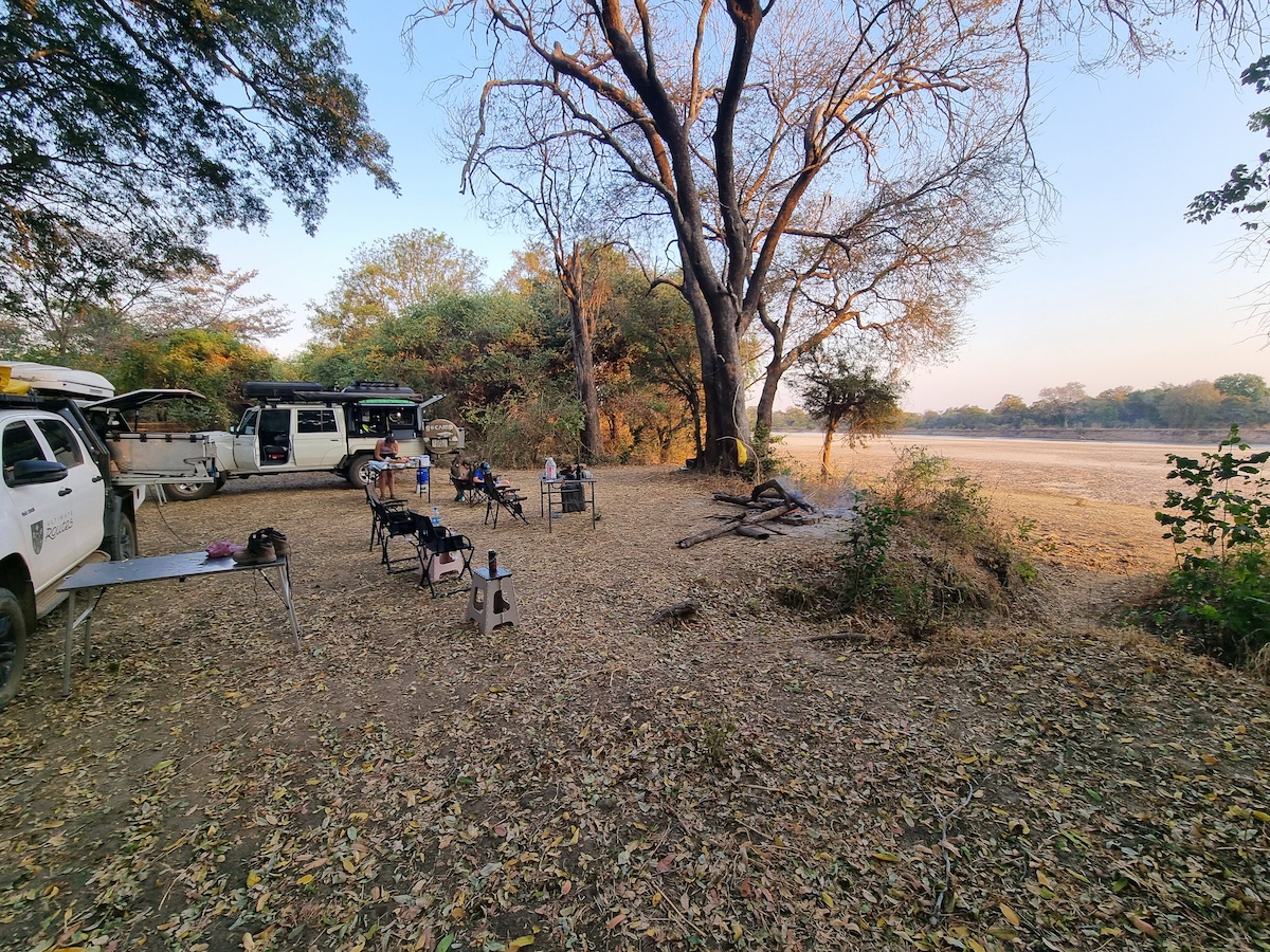 Two equipped 4x4s camped under trees beside a sandy riverbed